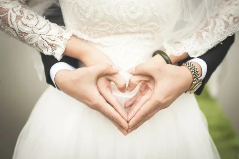 Two people in formal attire form a heart shape with their hands.