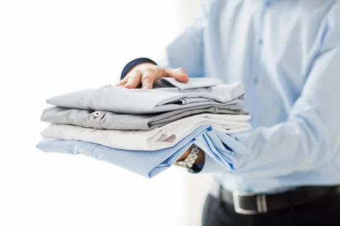 A person in a blue dress shirt holds a neatly folded stack of shirts in various colors, including white, grey, and blue.
