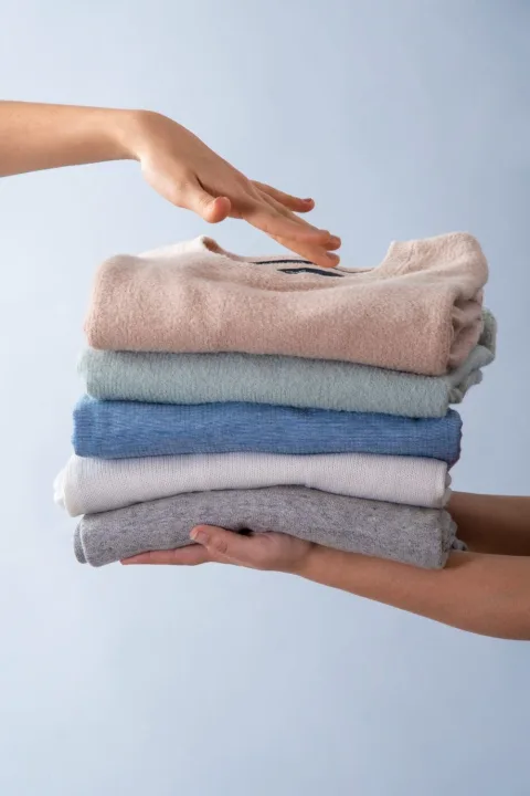 A person places a hand on top of a neatly folded stack of sweaters in different colors, held by another person against a light background.