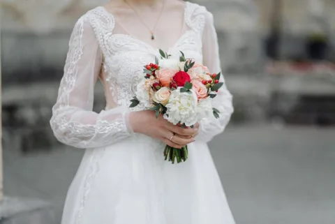 A bride in a white lace wedding dress holds a bouquet of mixed flowers, including white, pink, and red blooms, with greenery.