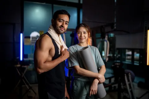 Two people in athletic clothing stand in a gym; one holds a towel over their shoulder, and the other holds a yoga mat.
