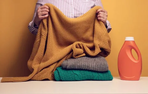 Person holding a mustard sweater, with folded gray and green sweaters on a table, alongside an orange detergent bottle, against a yellow background.