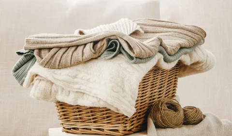 A wicker basket filled with folded knit sweaters in neutral colors, with two balls of yarn placed beside the basket.
