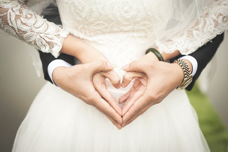 Two people in formal attire form a heart shape with their hands.