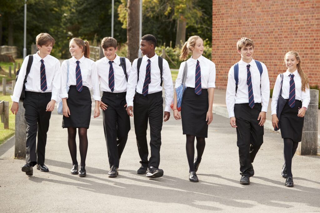 A group of students in school uniforms walk together outside, wearing white shirts, striped ties, and black pants or skirts.