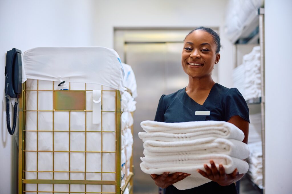 A woman in uniform stands in a laundry room holding a stack of white towels, with a towel cart and shelves in the background.