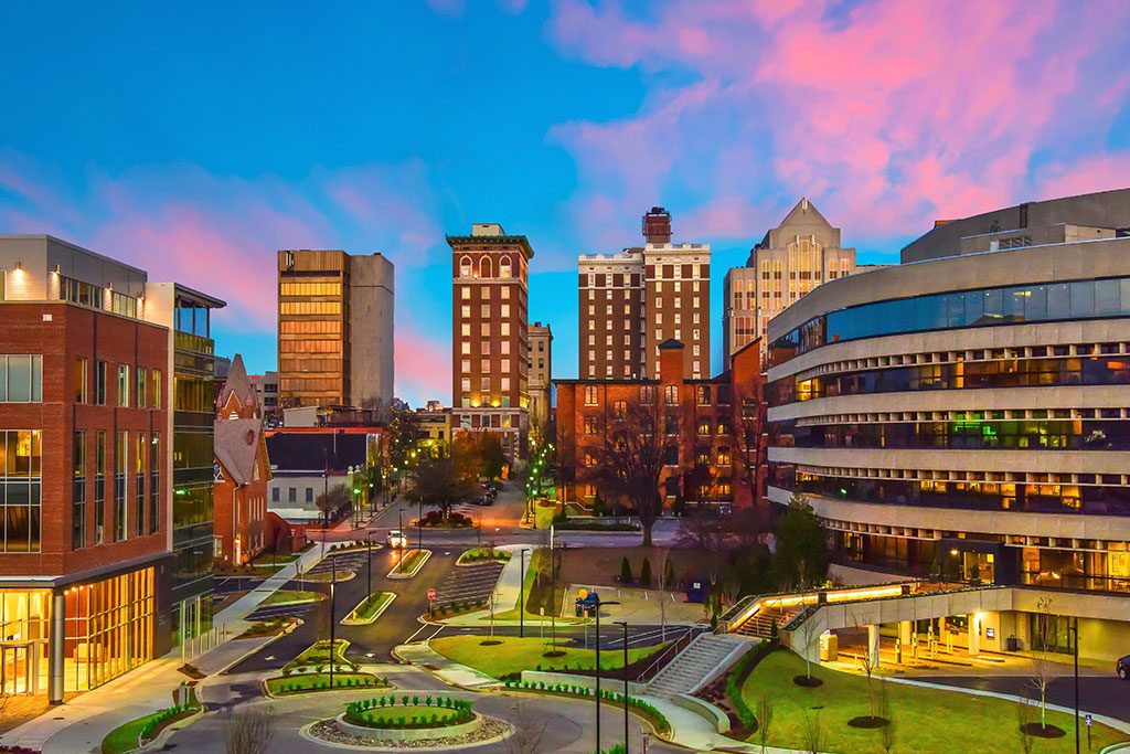 Cityscape at dusk with modern and historic buildings, including a curved glass structure on the right, under a sky with pink and blue hues. Streets and pathways are visible in the foreground.