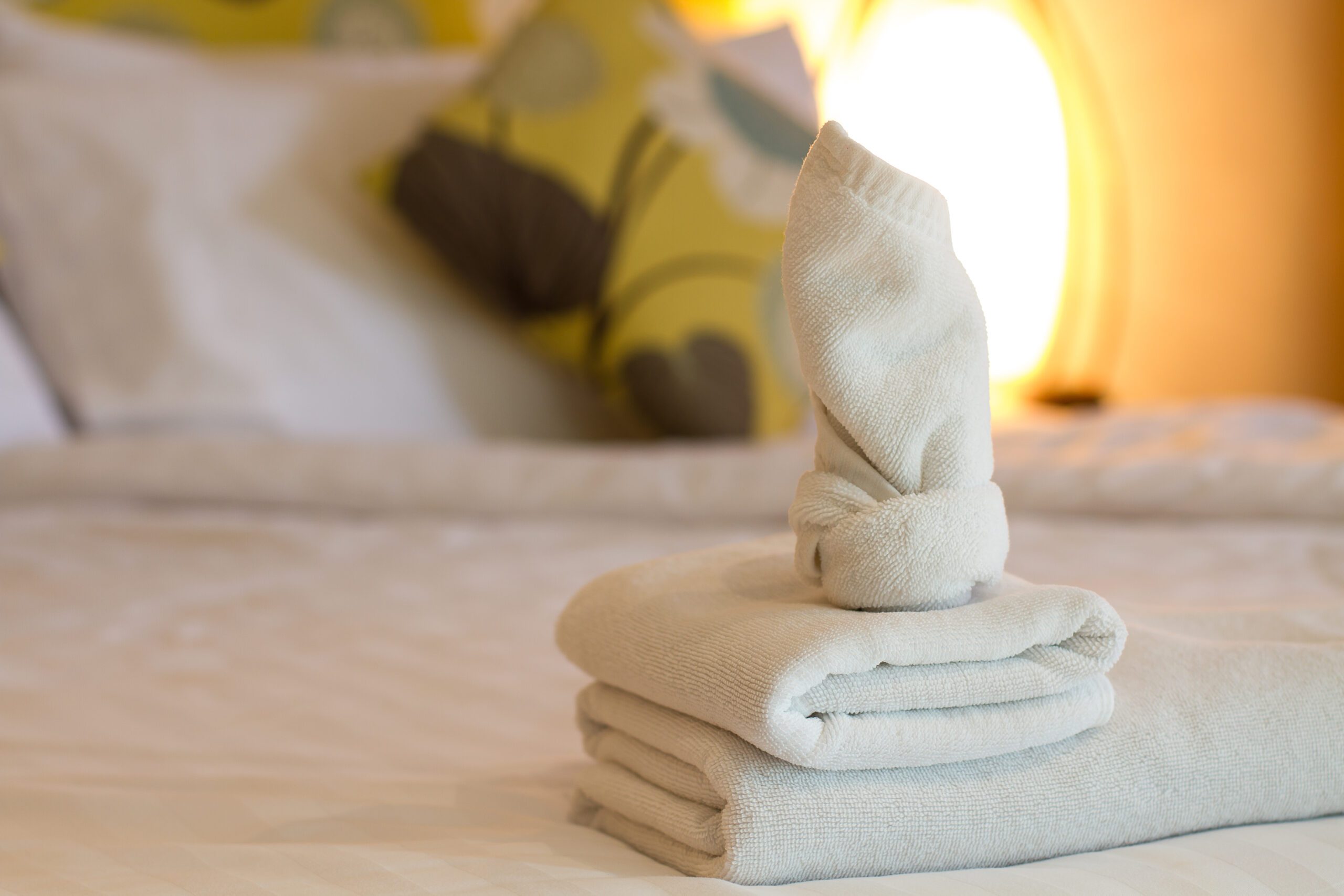 Folded towels with a towel sculpture on a bed, pillows and a lamp in the background.