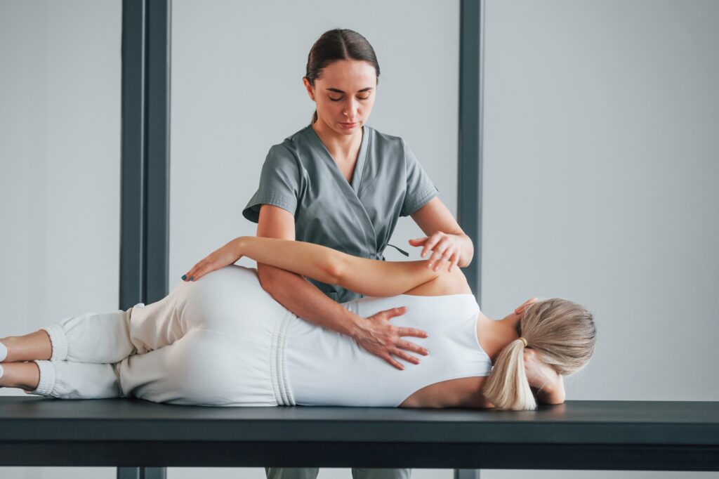 A therapist guides a patient through a side-lying stretch on a treatment table in a clinical setting.