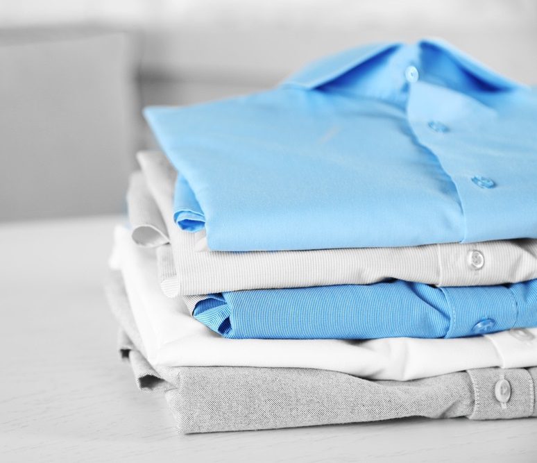 A neatly stacked pile of folded shirts, including light blue, gray, and white colors, placed on a white surface.