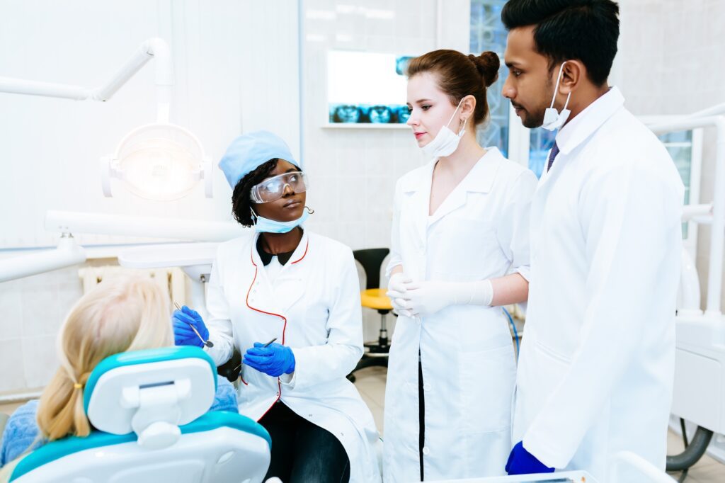 Three dental professionals in lab coats consult with a seated patient in a dental office.