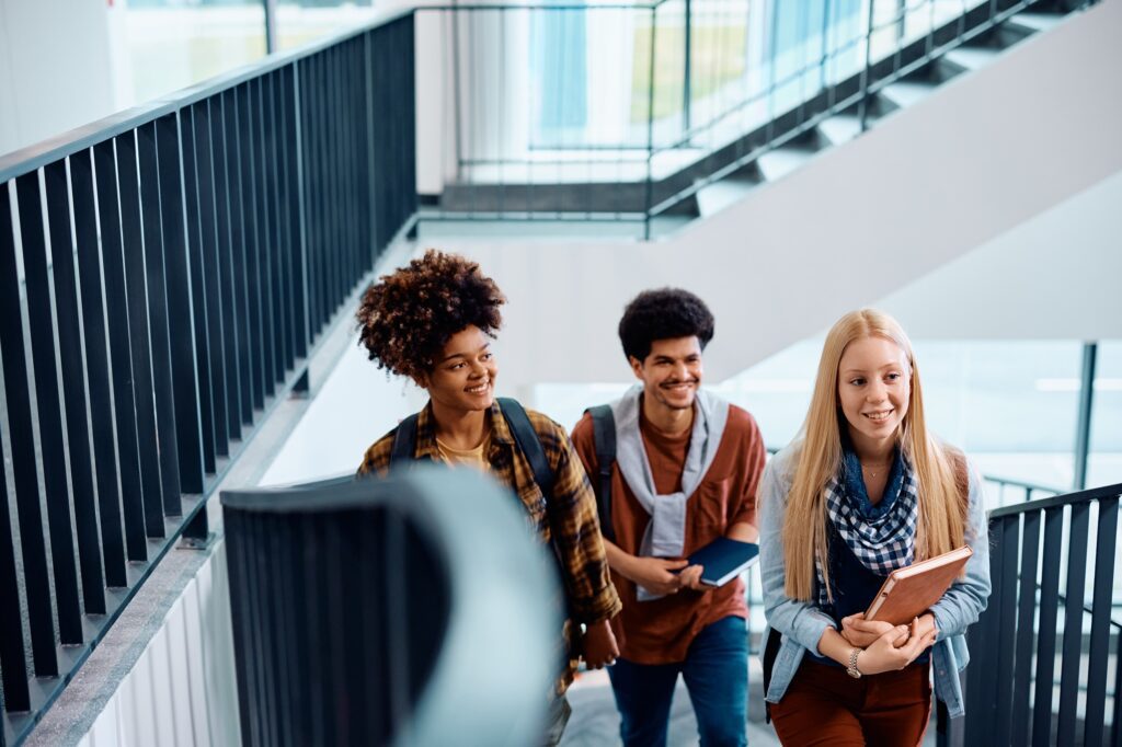 Three students walk up a staircase, each holding books and backpacks, smiling and engaged in conversation.