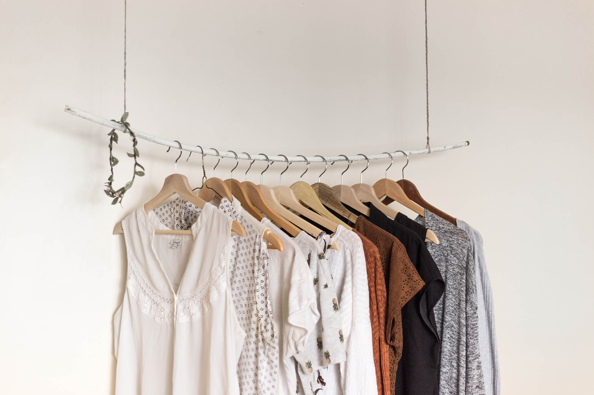 A minimalist clothing rack displays a selection of shirts in neutral tones, hung on wooden hangers against a plain white background.