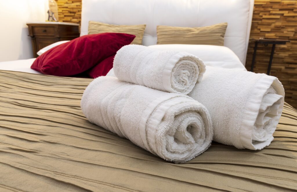 Three white rolled towels on a neatly made bed, with red and beige pillows in the background.