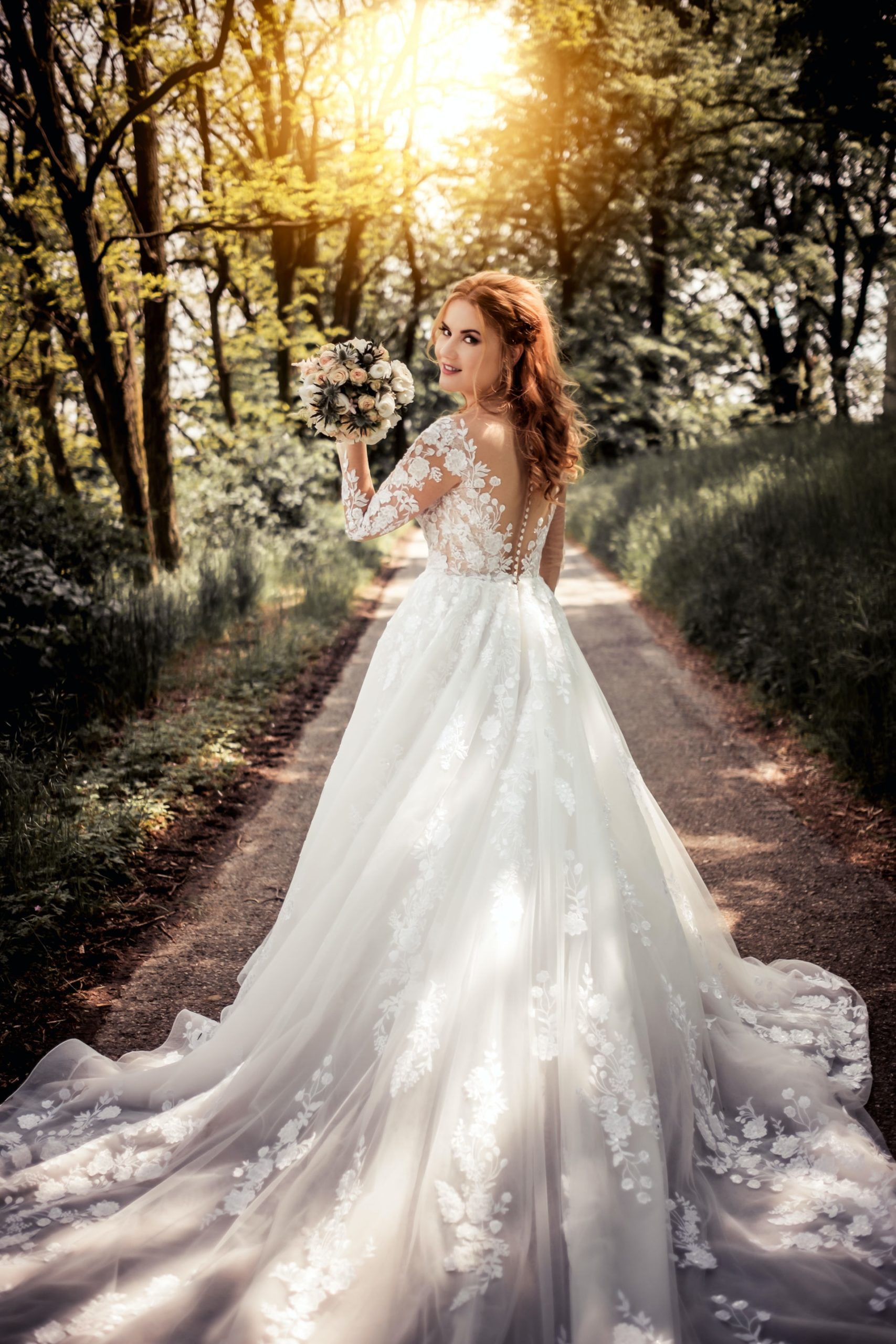 A woman in a lace wedding dress holds a bouquet and stands on a tree-lined path with sunlight filtering through.