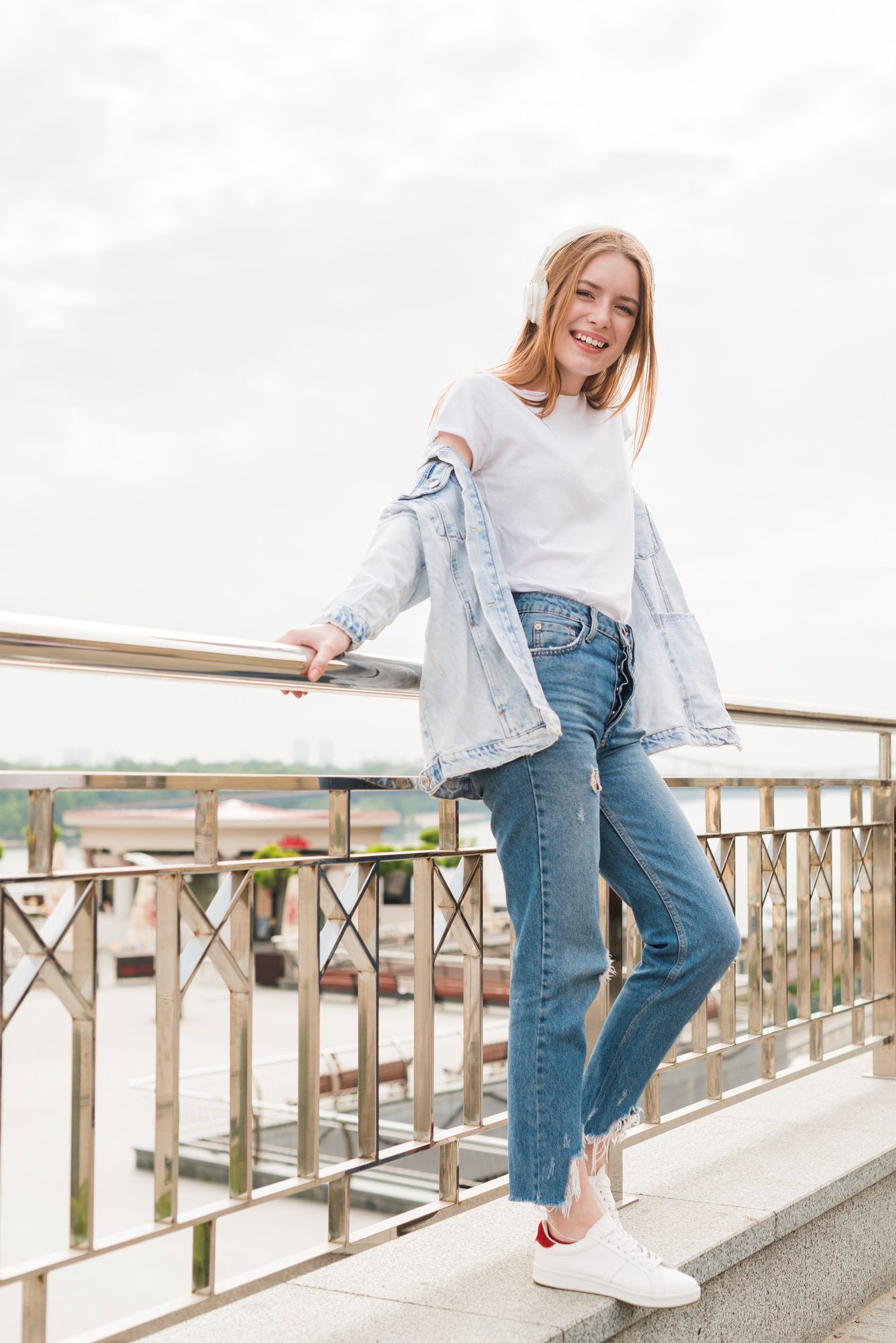 Person wearing headphones and casual clothing, standing and smiling on a bridge with a cityscape in the background.