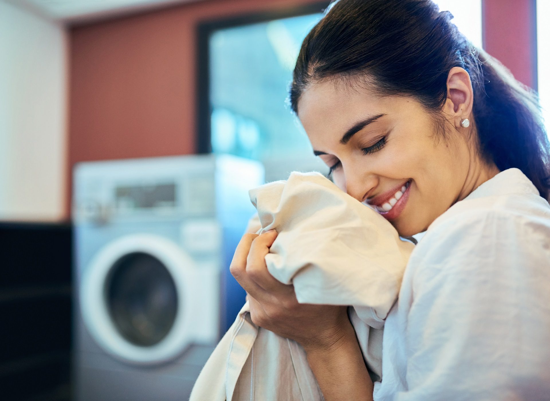 A woman smiles with her eyes closed while holding a piece of laundry near a washing machine.