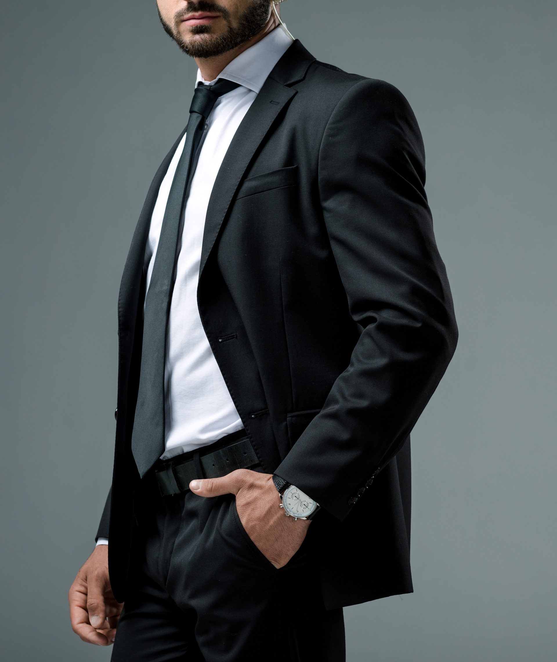 A man in a black suit and tie with a white shirt stands against a gray background, one hand in his pocket and the other resting casually at his side.