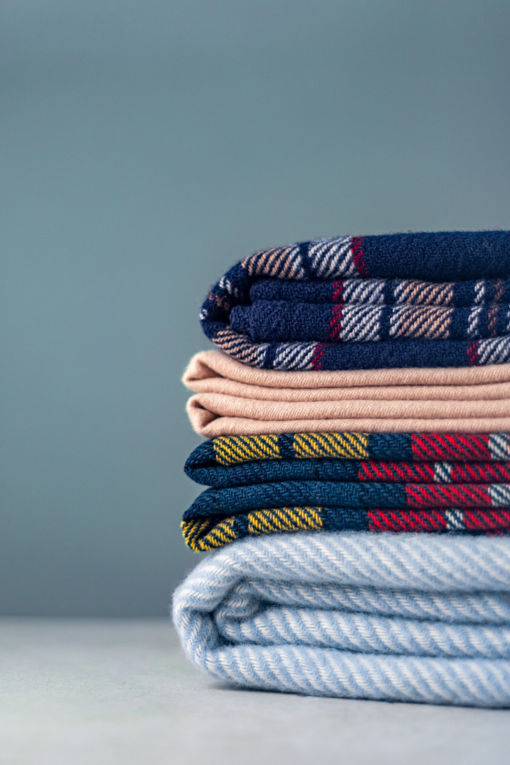 A neatly stacked pile of folded clothes with various patterns and colors, including plaid and striped designs, against a plain background.