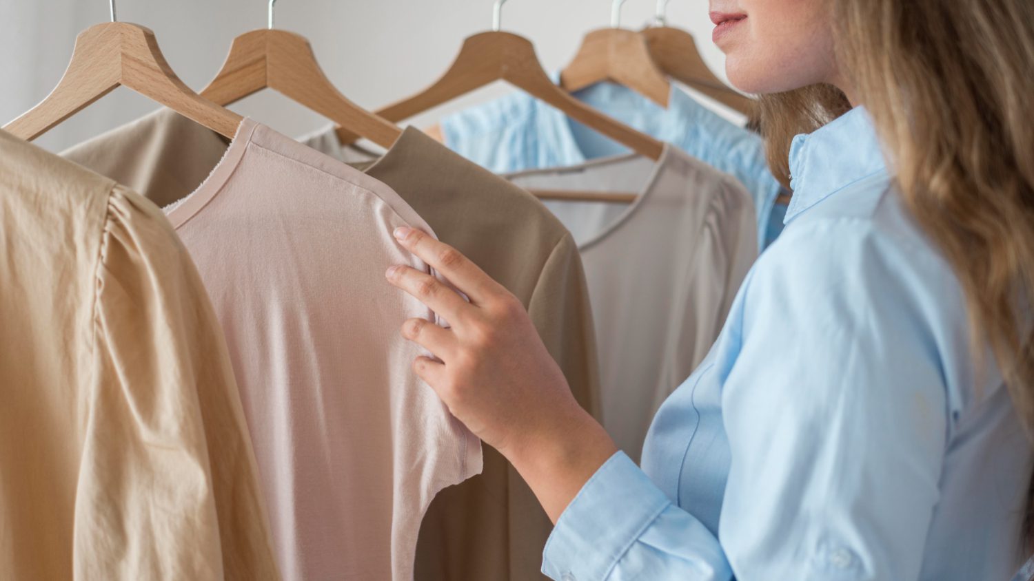 A person inspects clothing on wooden hangers, featuring various pastel-colored tops.