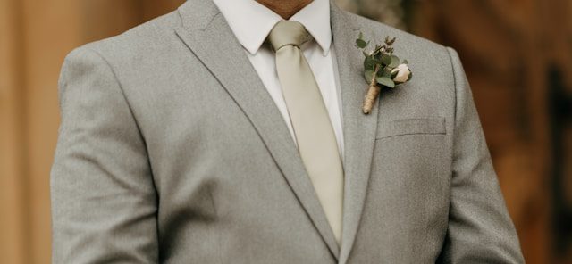 A man wearing a light gray suit, white shirt, and beige tie with a boutonniere pinned to his lapel.