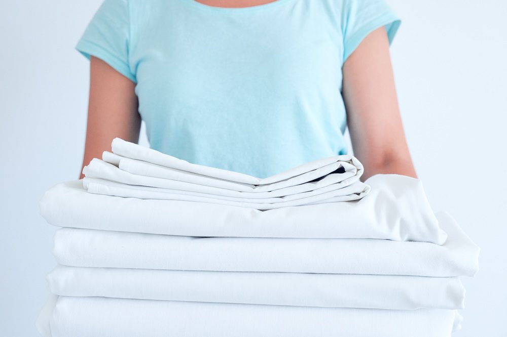 A person in a light blue shirt holding a stack of neatly folded white bed linens against a plain background.