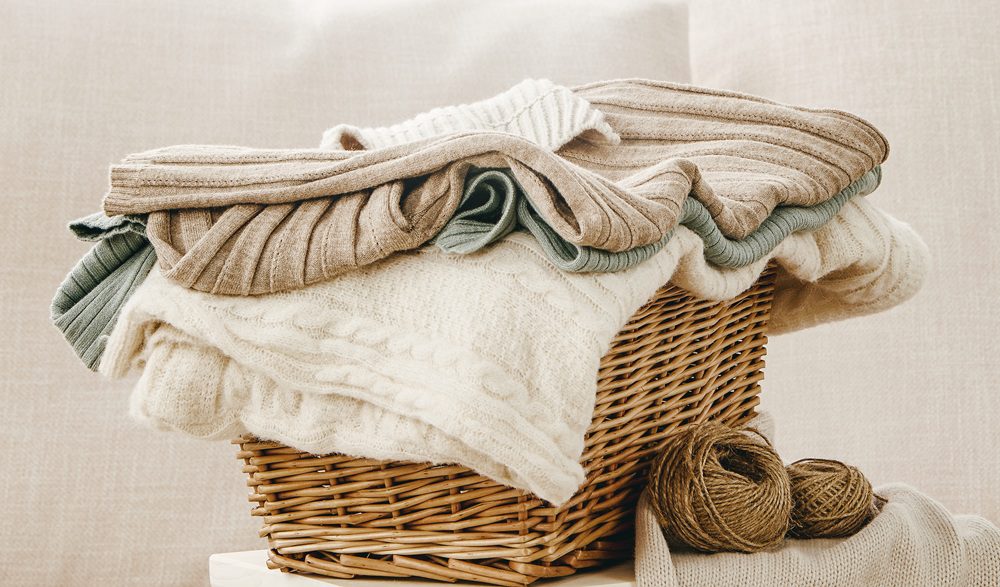 A wicker basket filled with folded knit sweaters in neutral colors, with two balls of yarn placed beside the basket.
