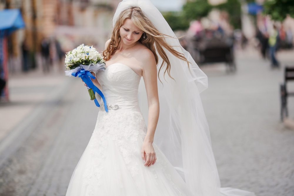 A bride in a white strapless wedding dress and veil holds a bouquet, standing on a cobblestone street outdoors.