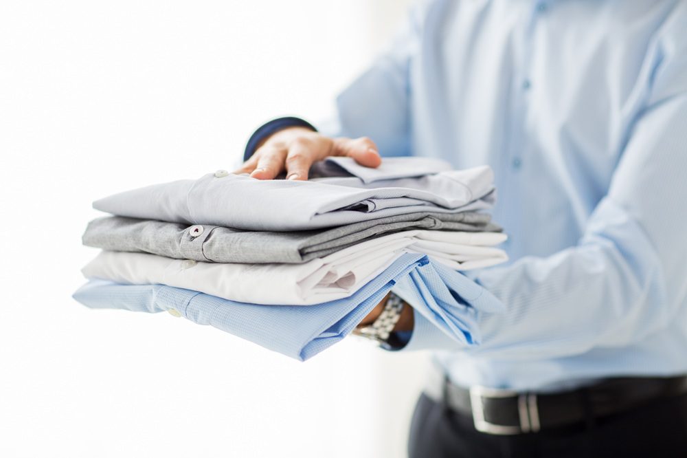 A person in a blue dress shirt holds a neatly folded stack of shirts in various colors, including white, grey, and blue.