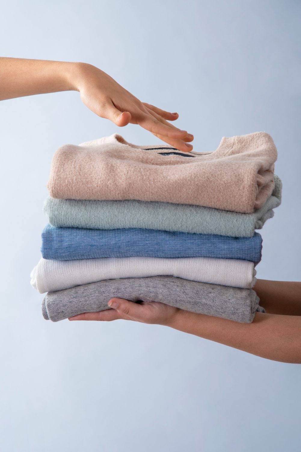 A person places a hand on top of a neatly folded stack of sweaters in different colors, held by another person against a light background.
