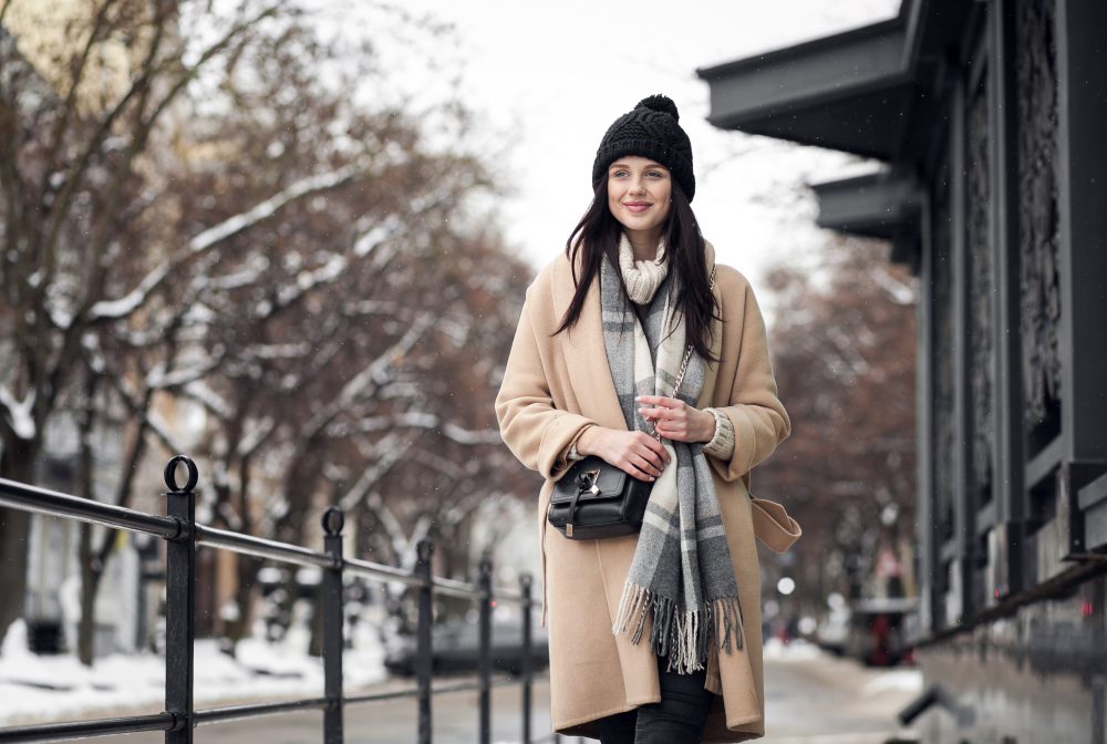 A woman in a beige coat and knit hat walks outside on a snowy city street, smiling and holding a cup.