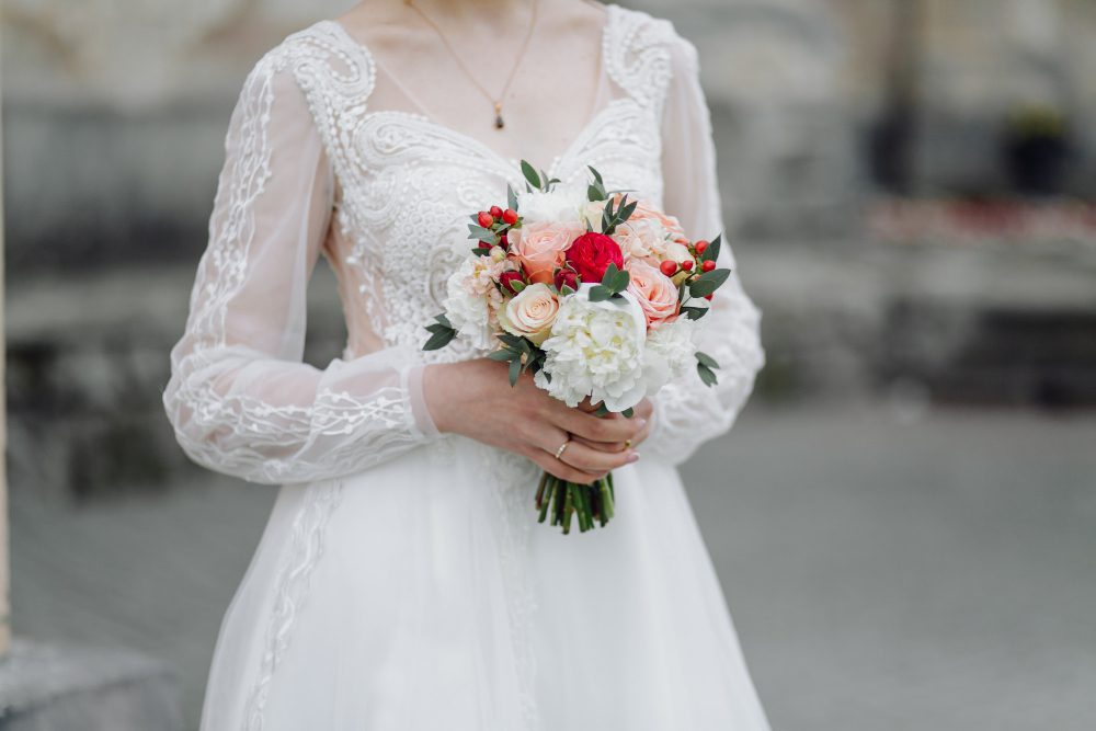 A bride in a white lace wedding dress holds a bouquet of mixed flowers, including white, pink, and red blooms, with greenery.